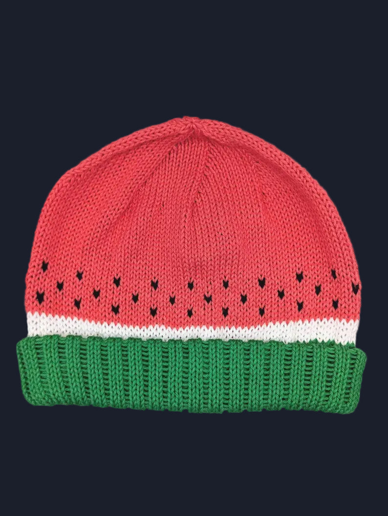 A knitted beanie hat designed to look like a watermelon slice, featuring a red top section with small black seed details, a white stripe, and a green ribbed bottom band.